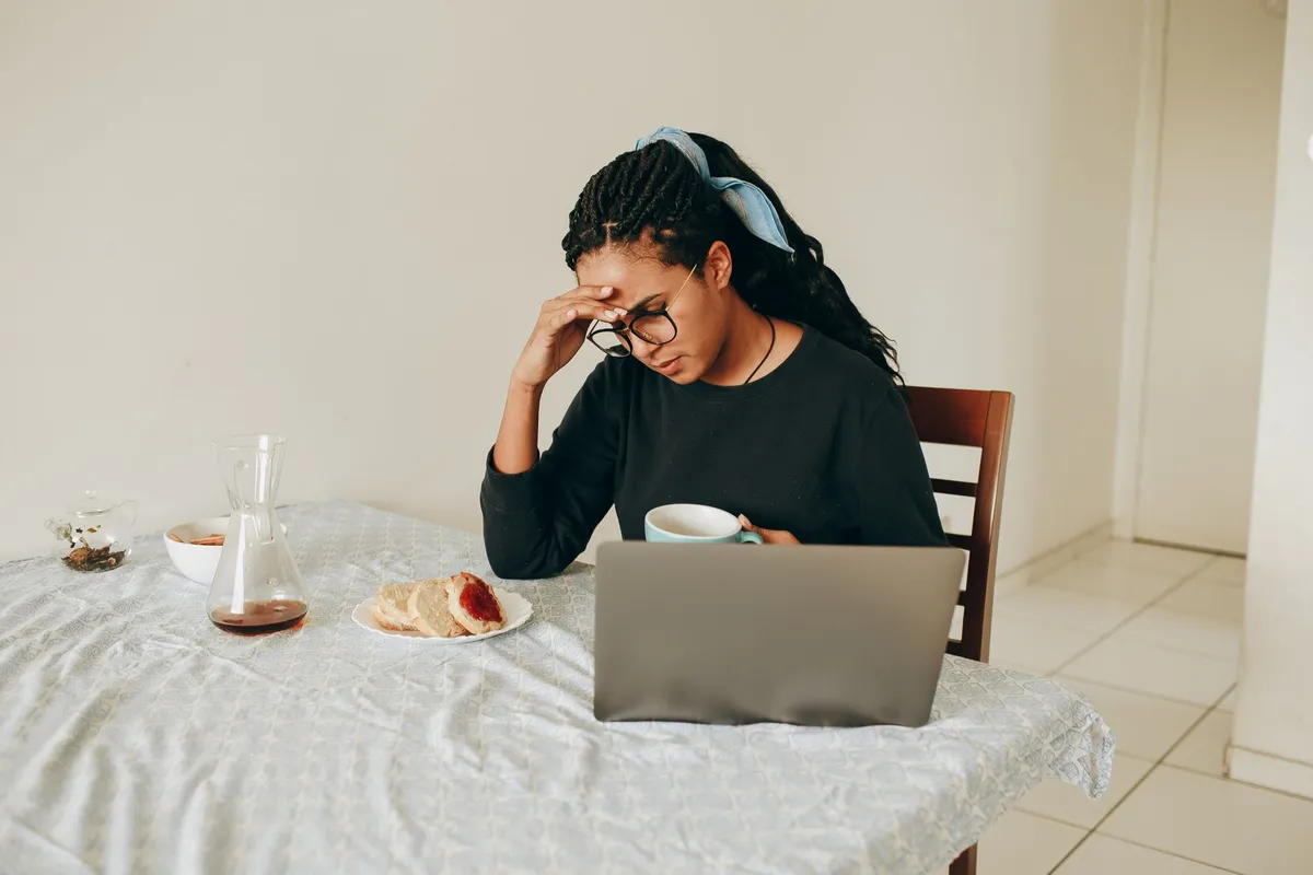 A woman sitting alone at a kitchen table in the morning with her hand against her forehead