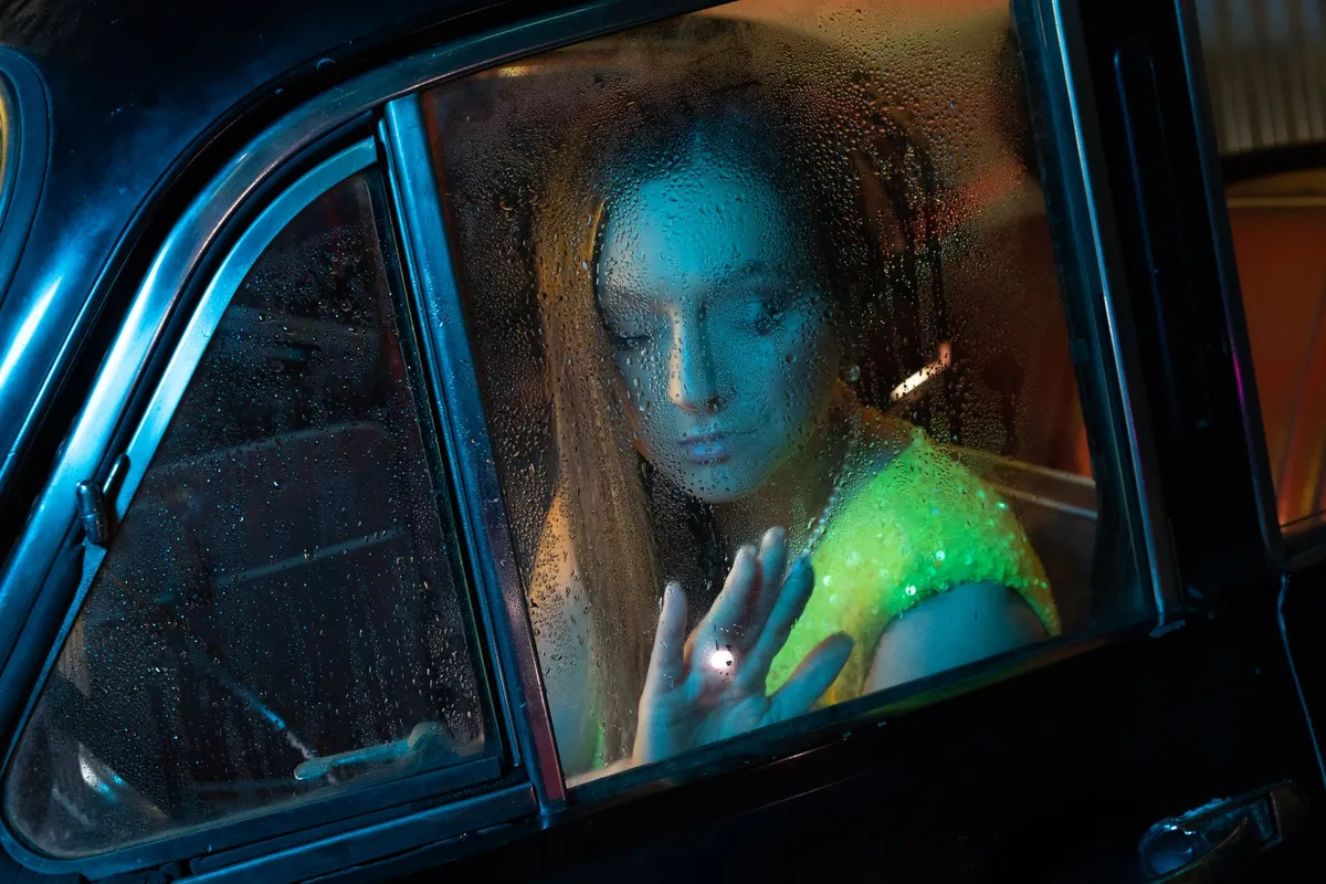A woman looking out a rain-streaked window alone, sitting with the signs of betrayal trauma