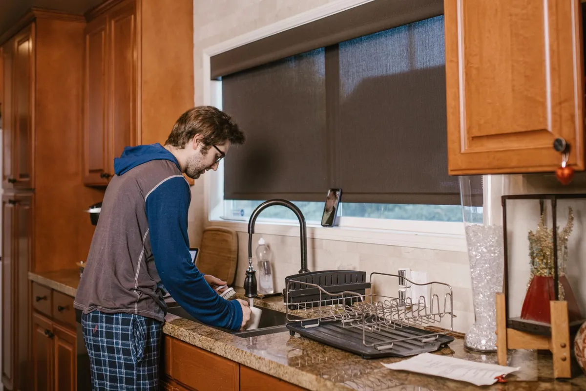 A man washing up at a kitchen sink with a phone propped on the windowsill