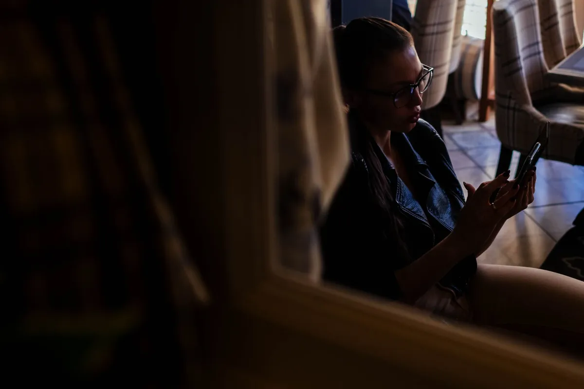 A woman sitting alone in low light looking at her phone, in the early stages of betrayal trauma
