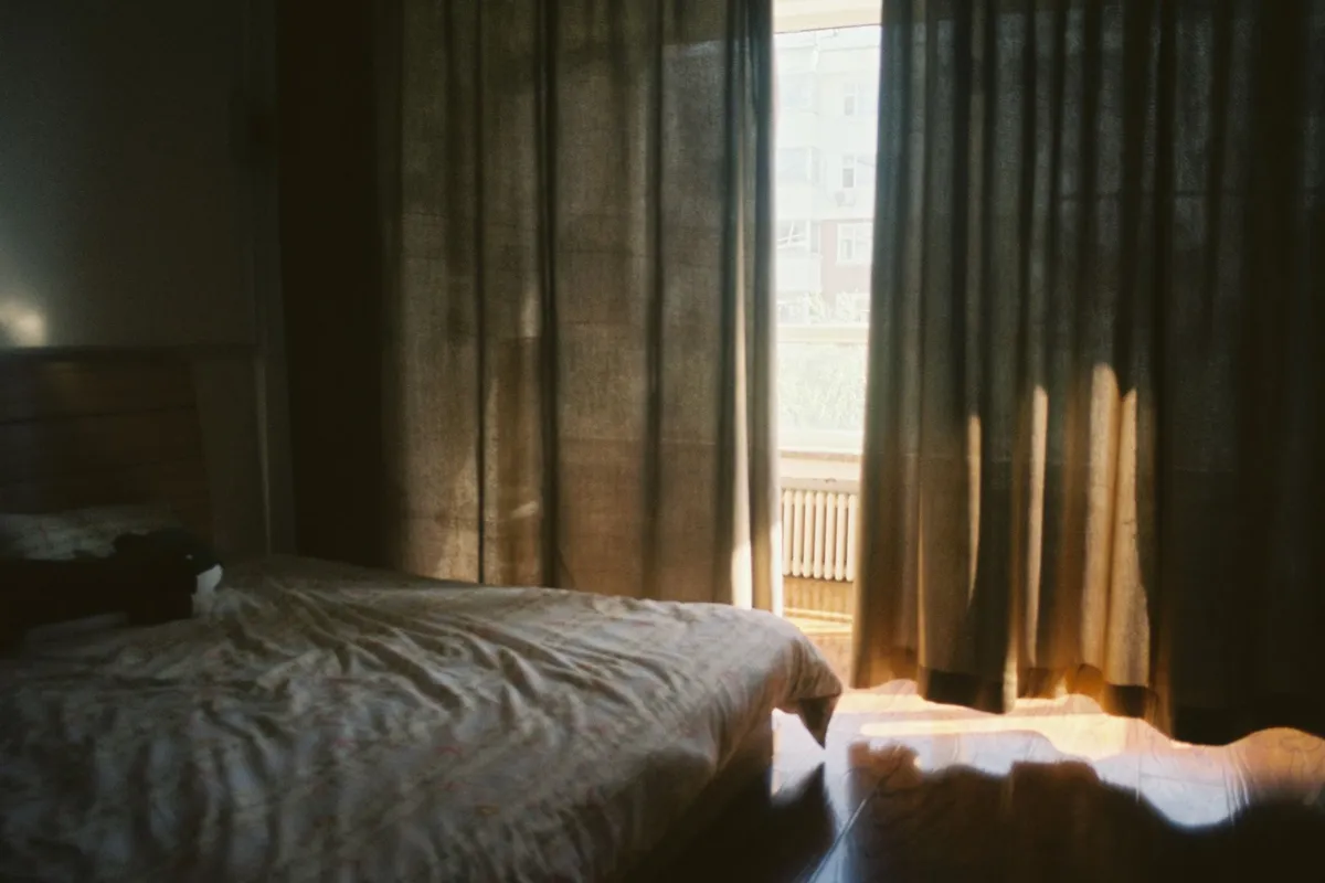 An empty half-made bed in a dim bedroom with light through the curtains