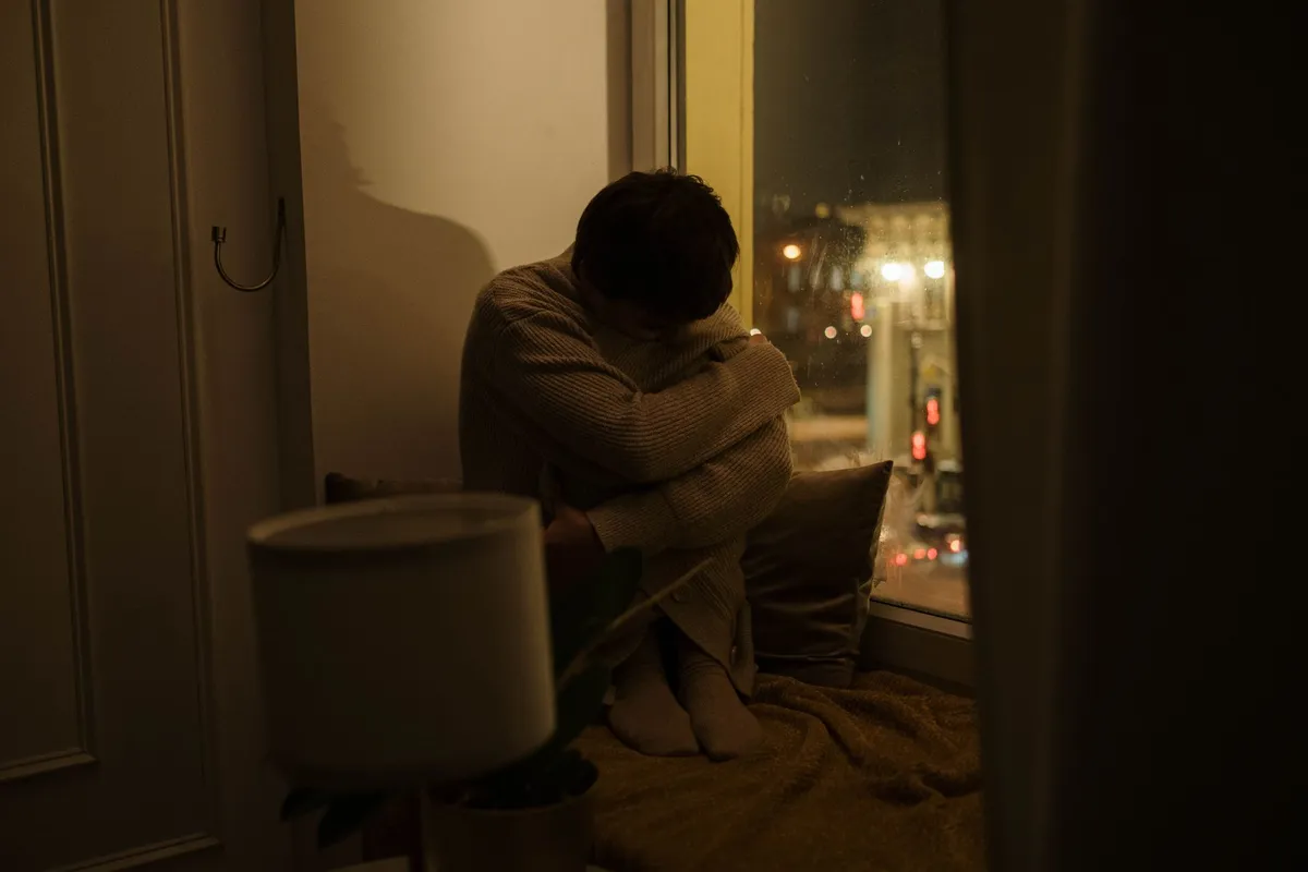 A man sitting curled up on a window seat at night with city lights behind him
