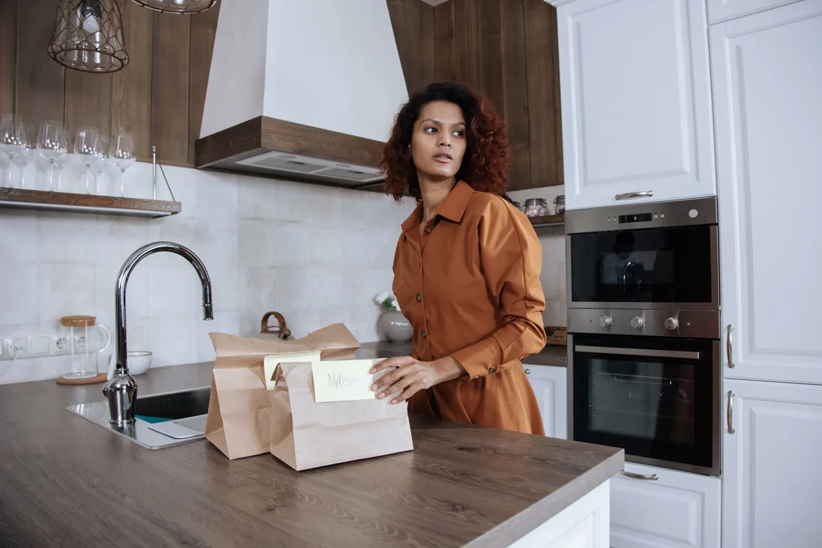 A woman standing at a kitchen counter with grocery bags, turning to look over her shoulder