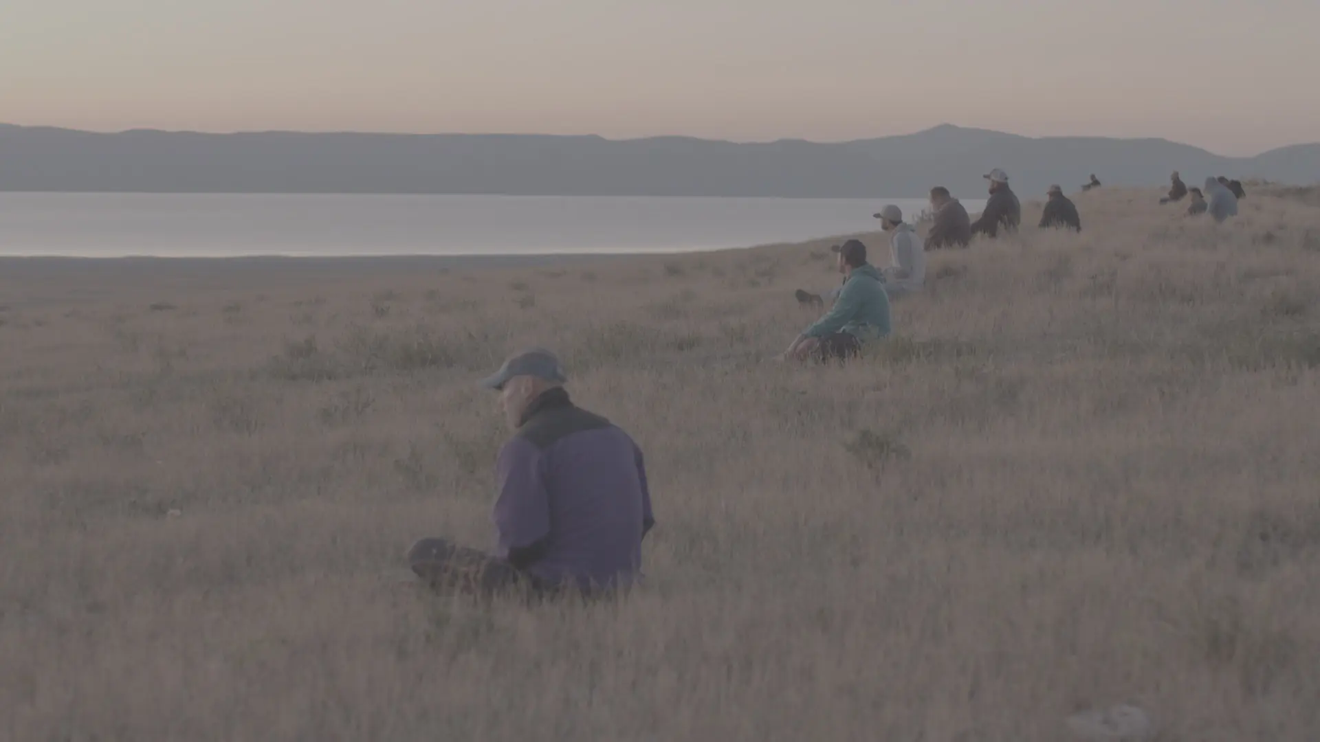 Men sitting apart in a golden field by a lake at dawn, taking time to reflect